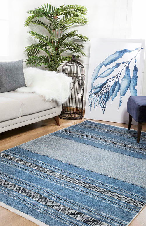 Load image 1 in gallery view. A modern living room featuring the Layered Lines - Washable Rug - JR1545 in blue patterns, ideal for high-traffic areas, paired with a light gray sofa, white faux fur throw, potted plant, birdcage, blue artwork, and dark blue stool on wood flooring.