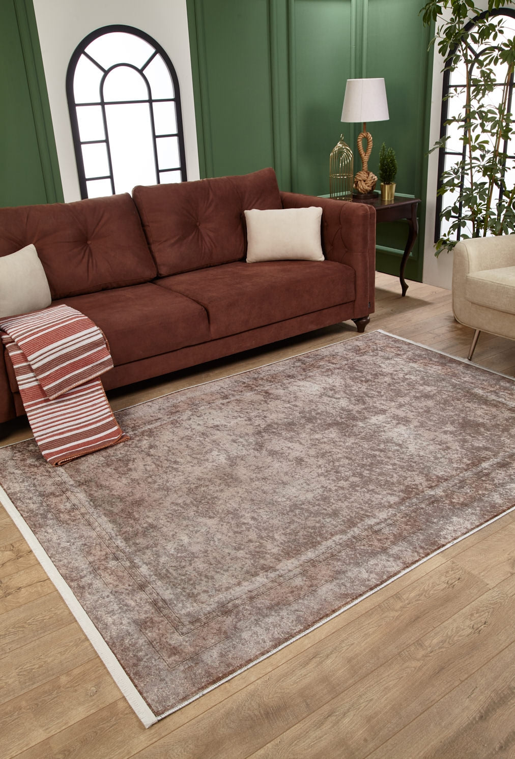 A cozy living room with a brown sofa, cream pillows, and a beige armchair features the Luxe Borderline - Washable Rug - JR1741—perfect for high-traffic areas. A side table with a lamp and decor sits against a green-paneled wall.