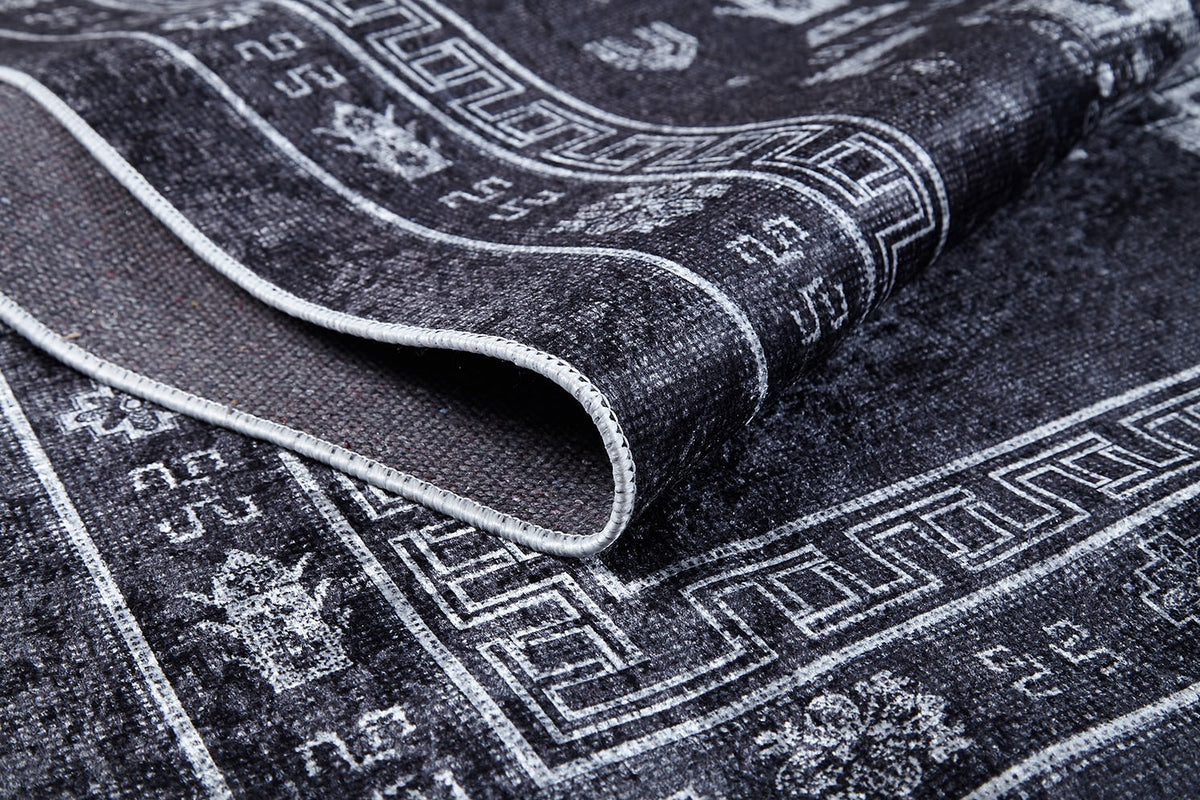 A close-up of the Majestic Symbol - Washable Rug (JR1892) in a dark tone, featuring intricate white geometric and floral designs, highlights its flat weave texture and detailed edge stitching with part of the rug folded over.
