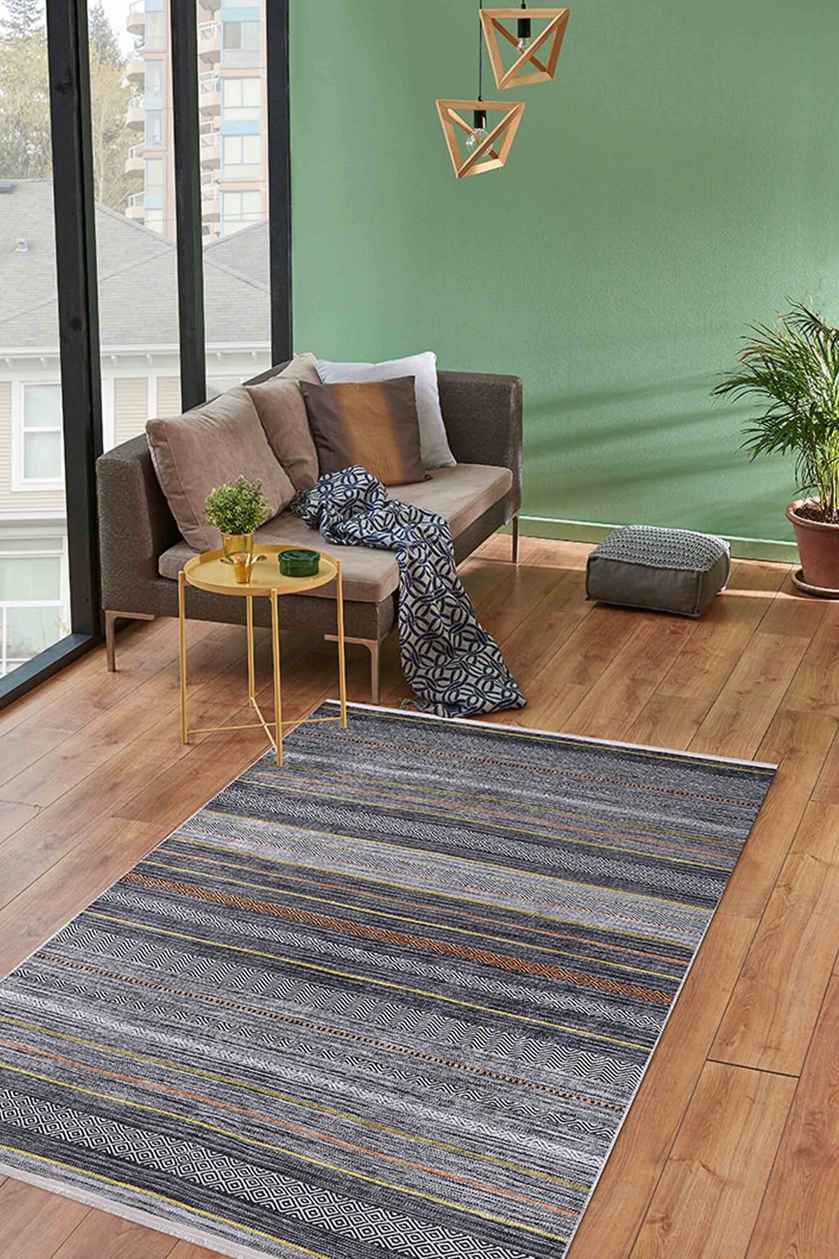A cozy living room features a small beige sofa, patterned throw blanket, yellow side table, Metro Lines - Washable Rug - JR1548 in gray-anthracite for high-traffic areas, potted plant, floor cushion, geometric hanging lights, green walls, and large windows.