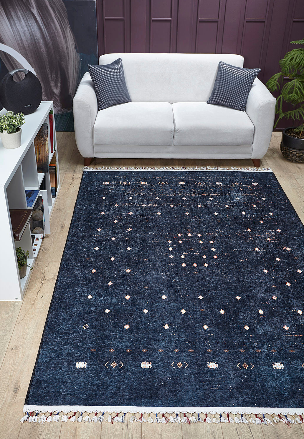 A modern living room with a light gray sofa, two dark pillows, a Midnight Majesty machine-washable rug (JRS5052) in dark blue pattern on wooden floor—perfect for high-traffic areas—a white shelf with books and plants, and a potted plant in the corner.