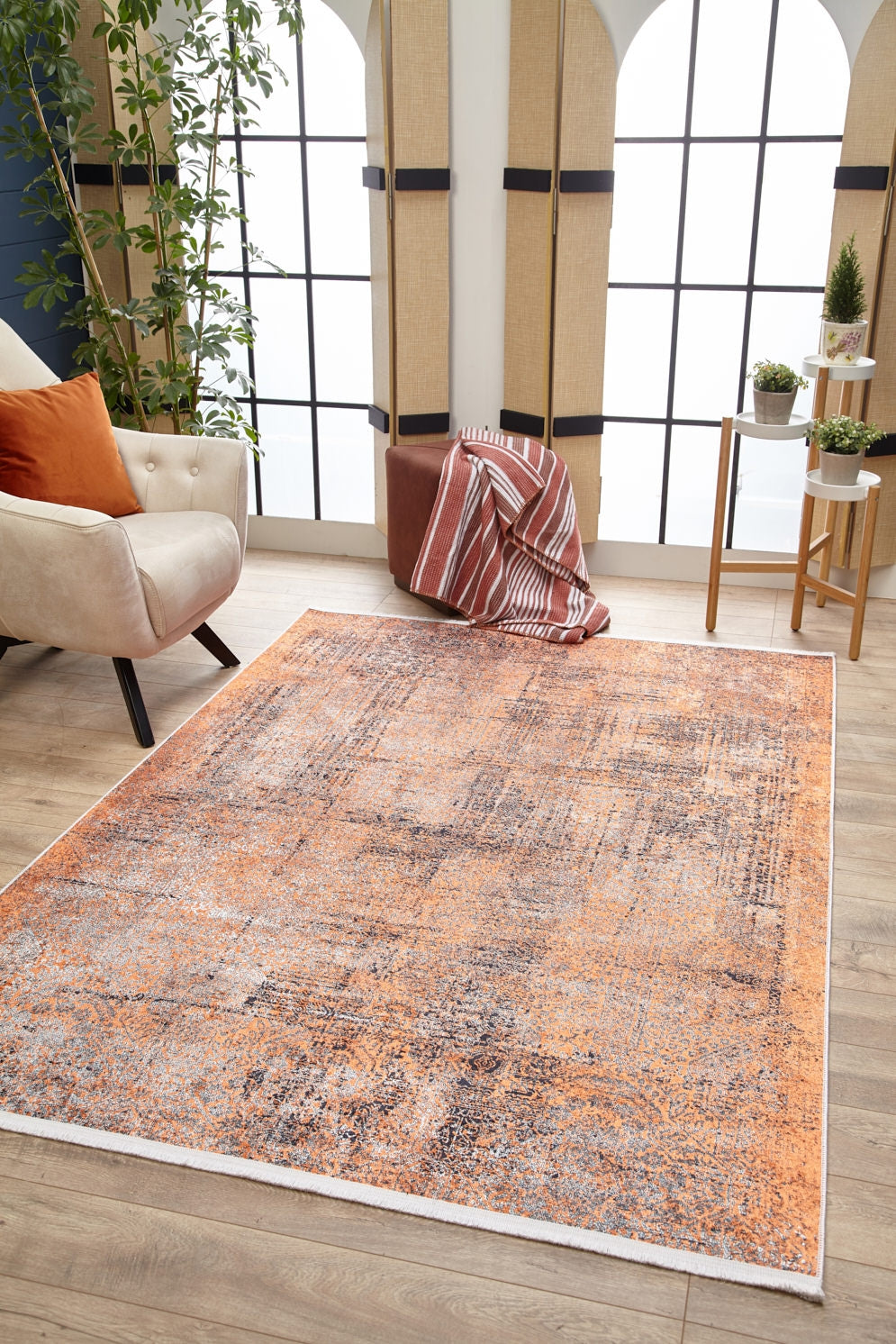 A living room with large grid windows, the Modern Mosaic - Machine-Washable Rug JR1846 in orange and gray for high-traffic areas, a beige armchair with orange pillows, brown ottoman with striped blanket, potted plants, and wood flooring.