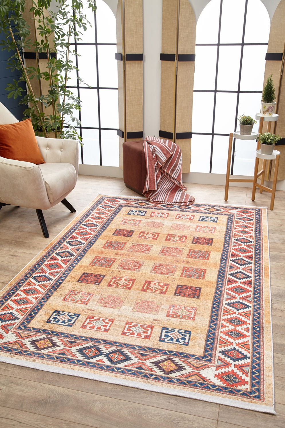A living room features the Navajo Nights - Washable Rug - JR1758 with geometric designs for high-traffic areas, a cream armchair with orange pillows, a plant, a brown ottoman with a striped blanket, and two small tables by tall windows.