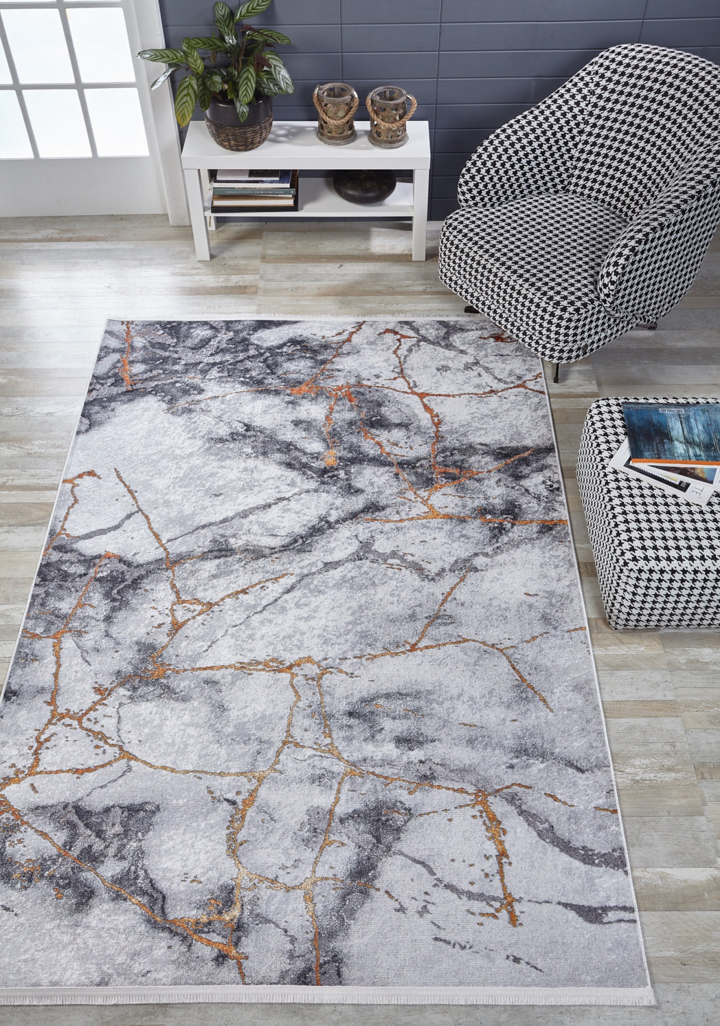 A modern living room features the Opulent Veins - Machine-Washable Rug JR1593 in gray-anthracite with gold veining, a black-and-white houndstooth armchair, ottoman, small white table with decor, plant, and light wood floors—ideal for high-traffic spaces.