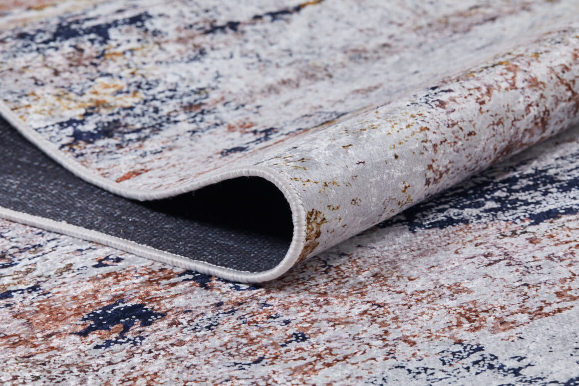 A close-up of the Painted Dreams - Washable Rug - JR432 showcases its abstract white, blue, brown, and gold patterns. One edge is lifted to reveal its thickness and dark backing—perfect for high-traffic areas.