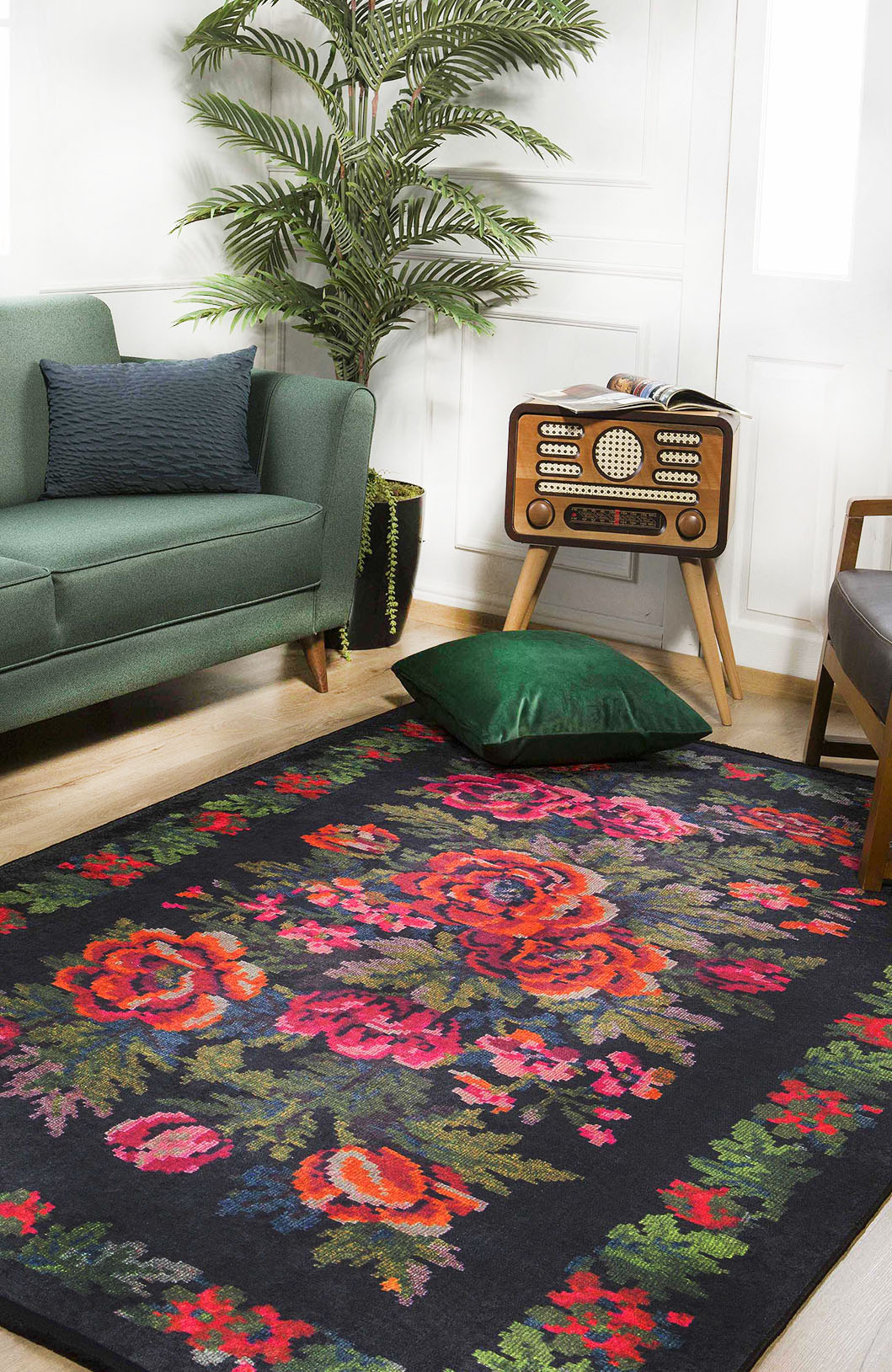 A living room with a green sofa, vintage radio on wooden legs, potted plant, and the JR294 Radiant Bloom washable rug featuring vibrant red and pink florals—perfect for high-traffic areas. A green velvet cushion rests on the rug.