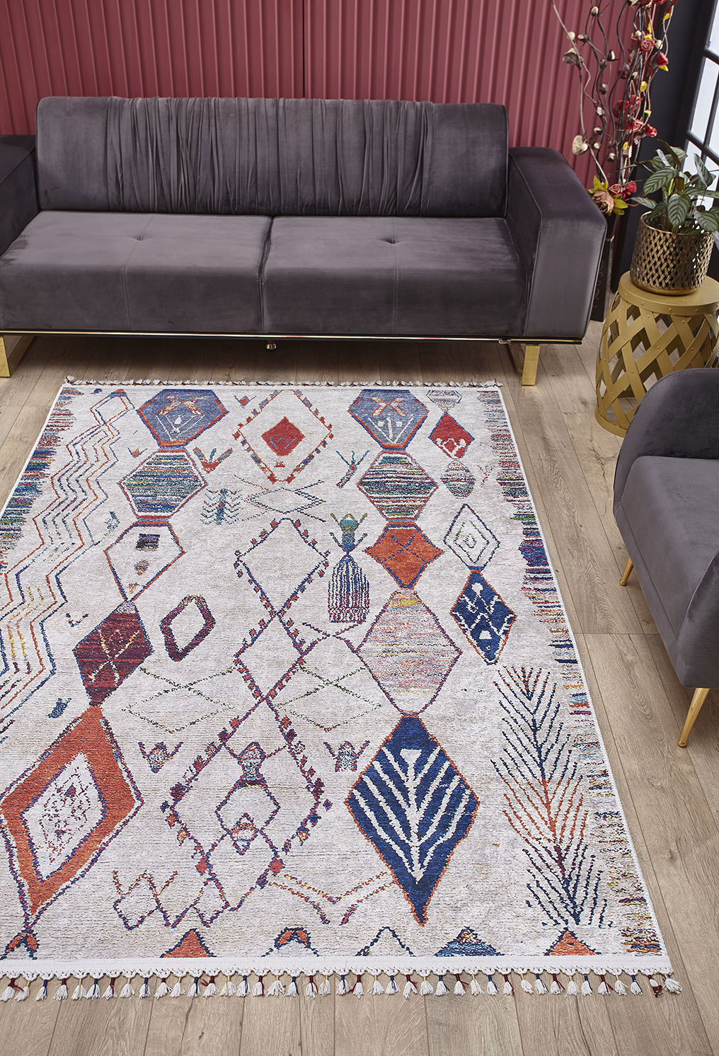 A modern living room features a gray velvet sofa, matching armchair, and the Rainbow Rhapsody - Washable Rug - JR5060 with a geometric pattern in red, blue, and orange on a light wood floor. Red vertical blinds and a decorative plant complete the background.