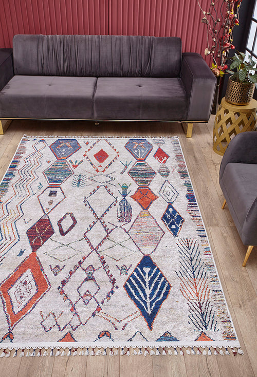 Bild 1 in Galerieansicht laden. A modern living room features a gray velvet sofa, matching armchair, and the Rainbow Rhapsody - Washable Rug - JR5060 with a geometric pattern in red, blue, and orange on a light wood floor. Red vertical blinds and a decorative plant complete the background.
