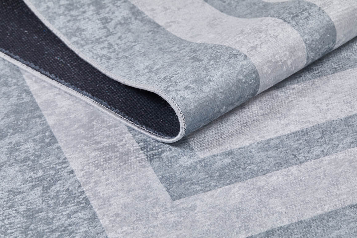 A close-up of Refined Edge - Washable Rug - JR573 in modern gray-anthracite with geometric patterns, partially folded to show its black non-slip backing—durable and ideal for high-traffic areas.