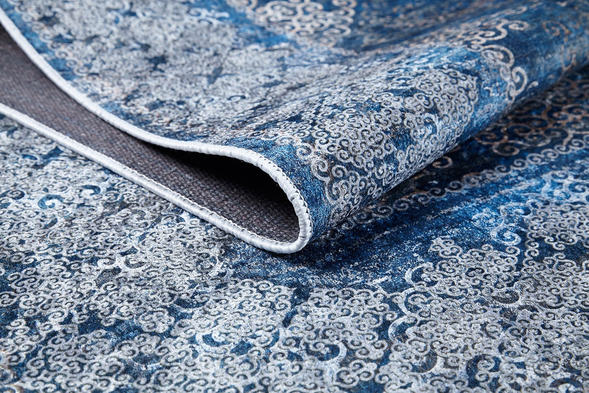 A close-up of the Regal Insignia - Washable Rug - JR1786 in blue, featuring intricate swirling designs and a folded edge revealing the underside—ideal for high-traffic areas.