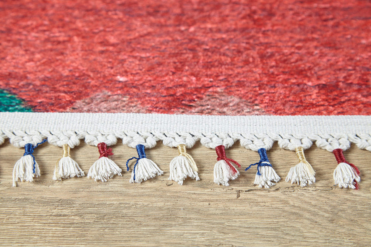 Close-up of the Rosy Blush - Machine-Washable Rug (JRS5067): red rug edge with cream tassels, some tied with red, blue, or yellow thread. Its vibrant red body features a hint of green in one corner and rests on a wooden floor.