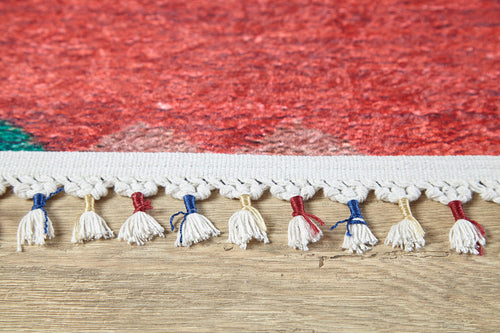Load image 1 in gallery view. Close-up of the Rosy Blush - Machine-Washable Rug (JRS5067): red rug edge with cream tassels, some tied with red, blue, or yellow thread. Its vibrant red body features a hint of green in one corner and rests on a wooden floor.