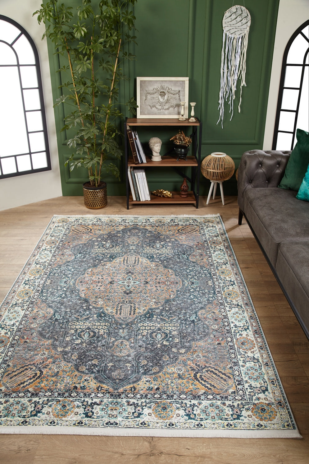 An elegant living room featuring the Royal Sultan - Washable Rug - JR1970 in blue, a gray tufted sofa with green pillows, art on a green wall, books on a small shelf, a plant, and black-framed windows—great for allergy-sensitive homes.