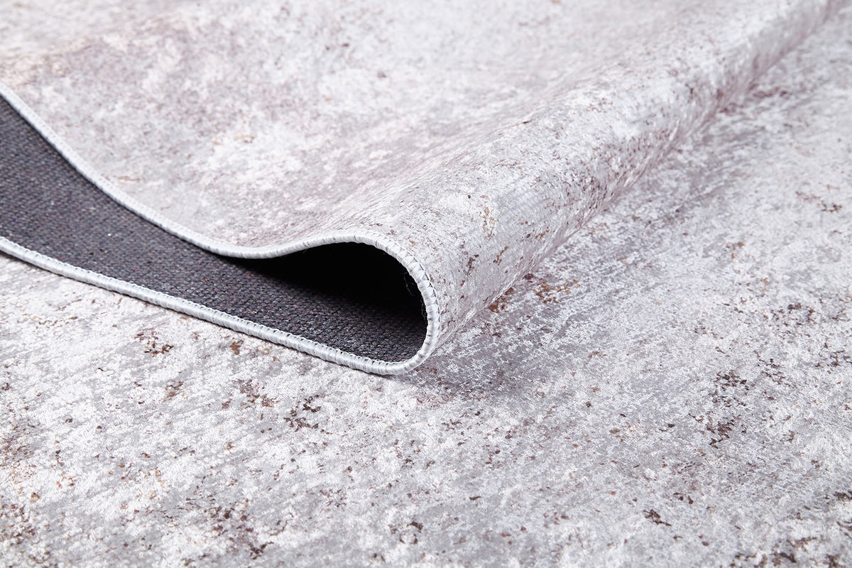 A close-up of the Rustic Trails - Washable Rug - JR1736 shows its textured light gray-anthracite surface and curled edge, revealing a dark non-slip backing—perfect for high-traffic areas.