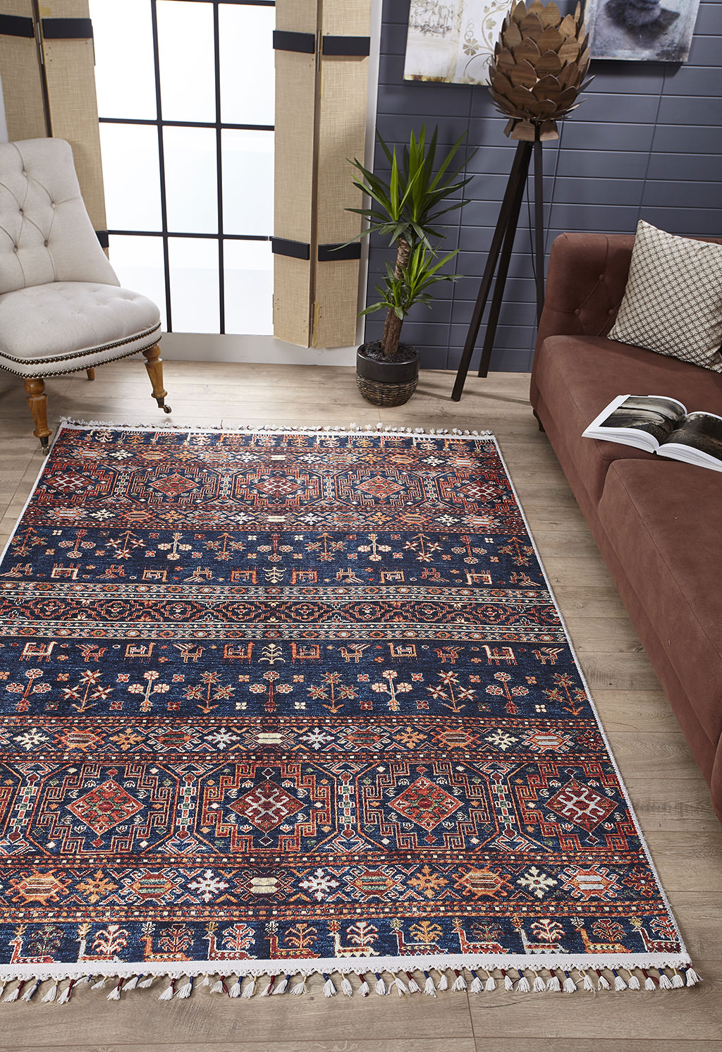 A living room featuring the Sahara Sunrise - Washable Rug - JR5091, ideal for high-traffic areas, a brown couch with an open book, a beige tufted chair, a potted plant, and large windows letting in natural light.