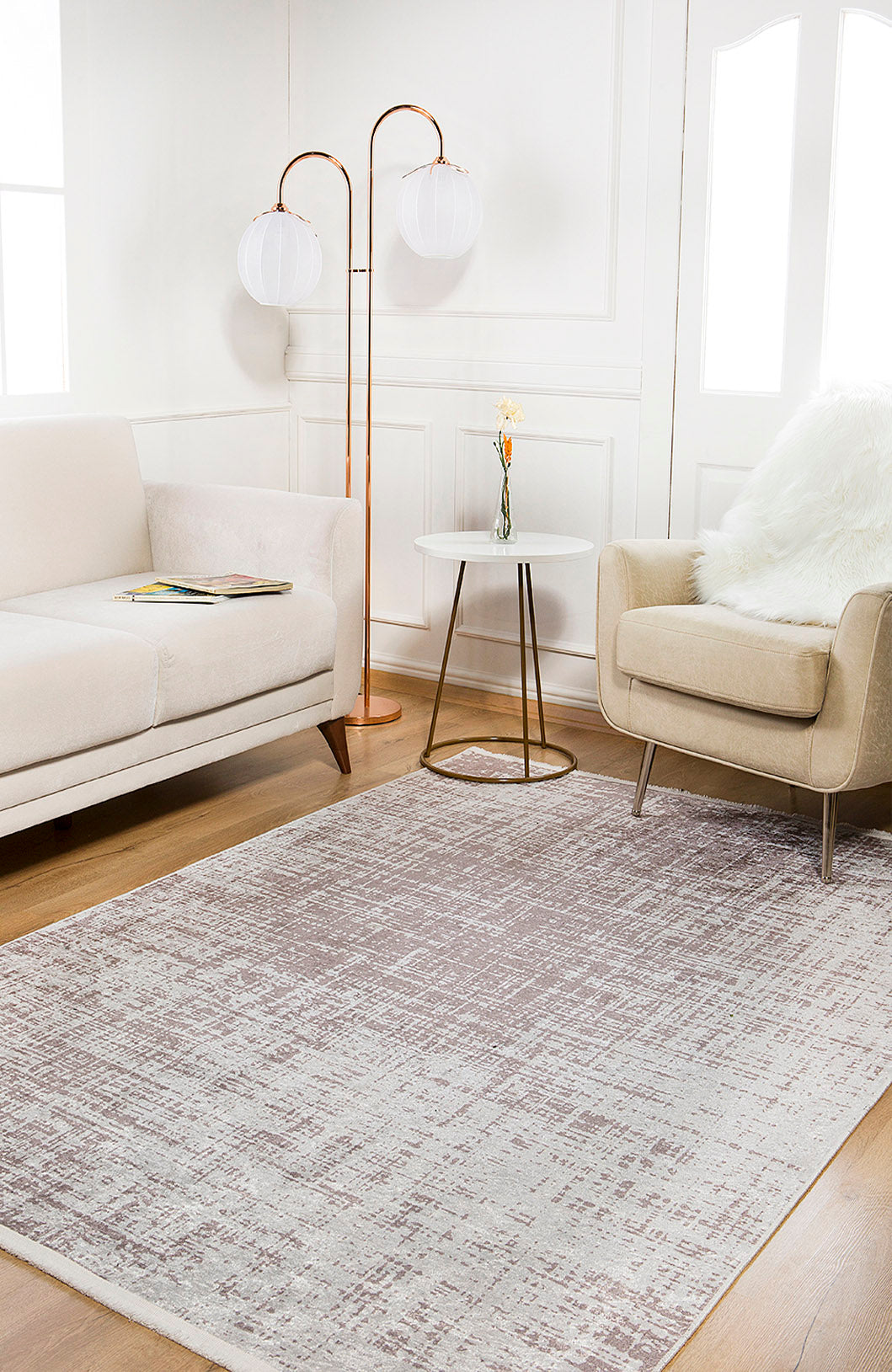 A modern living room features a beige sofa, tan armchair with a white throw, round side table with a small vase, globe floor lamp, and the Sea Spray - Washable Rug - JR847 in cream and beige on wooden flooring.