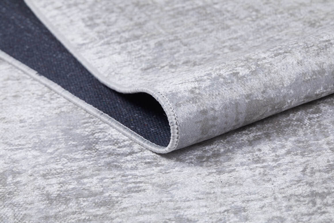 A close-up of the Sea Spray - Washable Rug - JR847 shows its light gray, flat weave and slightly shiny surface, with a corner curled to reveal the dark, non-slip backing. This washable rug blends style and practicality for any space.