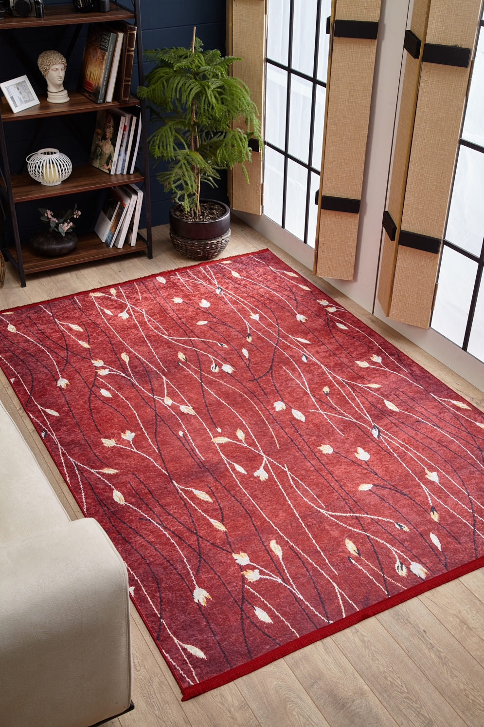 A living room with the Secret Garden - Washable Rug - JR1656, ideal for high-traffic spaces, showcases a white and beige floral vine pattern on red, paired with light wood flooring, a beige couch, potted plant, bookshelf, and bamboo-shaded windows.