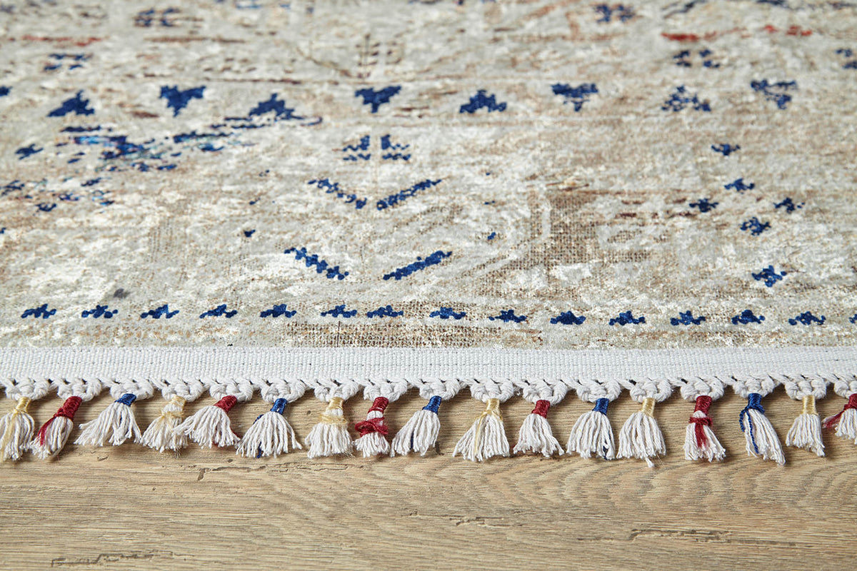 A close-up of the Shangri-La - Washable Rug - JR5042 in cream and beige with blue patterns and a red, blue, and white tasseled fringe, placed on a wooden floor—ideal for allergy-sensitive homes.