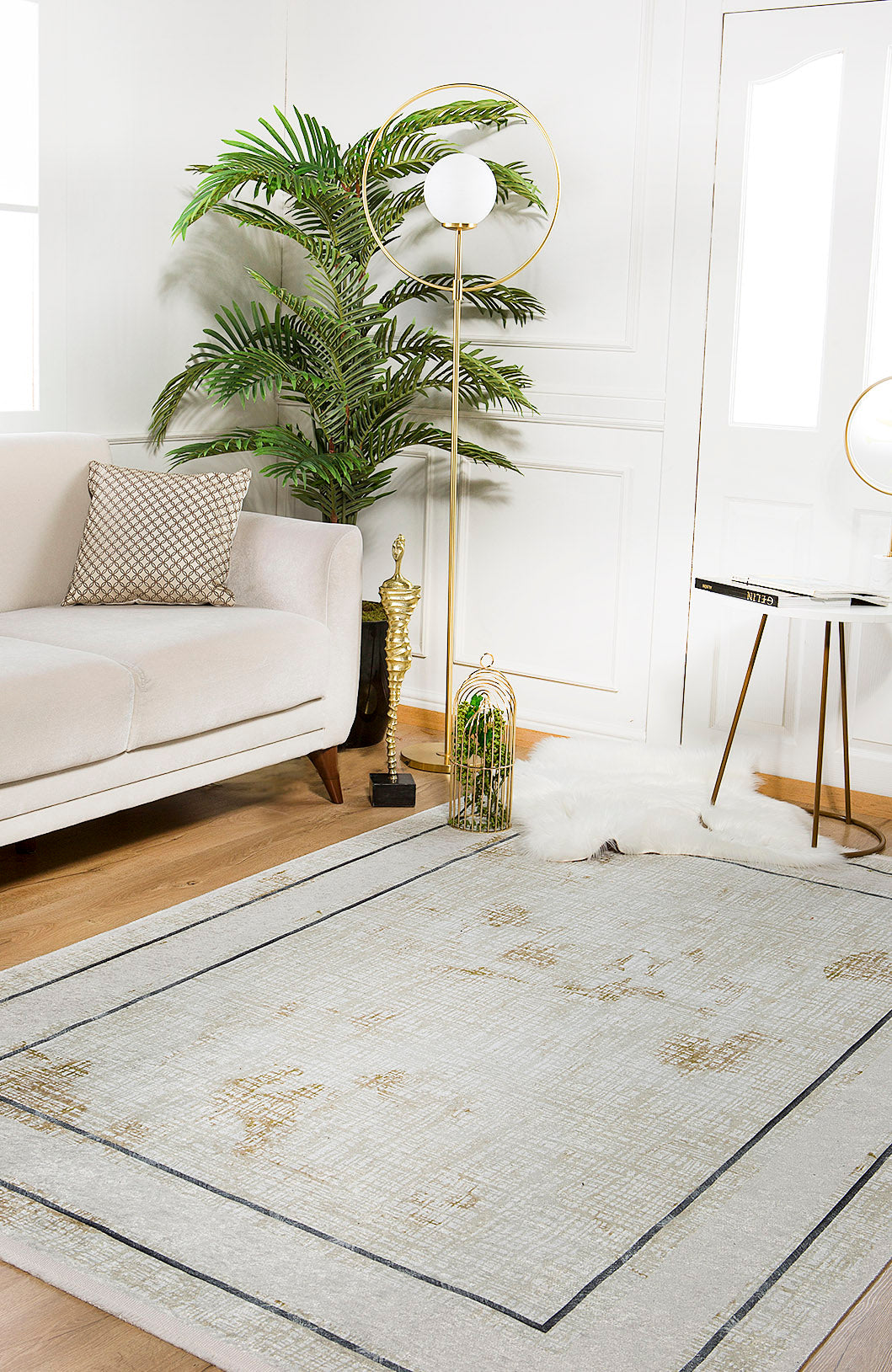 A modern living room featuring a light beige sofa, patterned pillow, potted plant, gold floor lamp, round side table, allergy-sensitive cream and beige rug, white and gold birdcage decor, and the Sleek and Contemporary - Washable Rug - JR1365 on wooden floors.