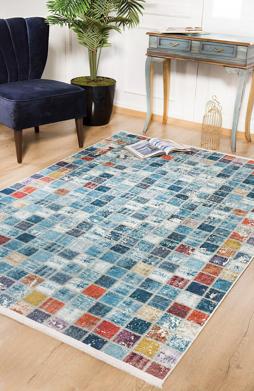 The Square Serenity - Washable Rug (JR1255), featuring blue, red, orange, and cream squares, is perfect for high-traffic areas and pairs well with wooden floors, a navy chair, blue desk, and indoor plants.