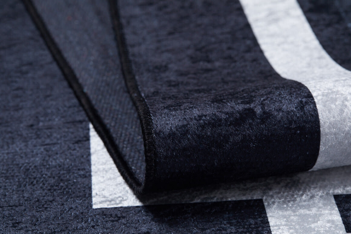 Close-up of the Subtle Border - Washable Rug - JR597 in black with a white geometric pattern; its edge is lifted, showing the soft, thick, and washable surface—ideal for high-traffic spaces.