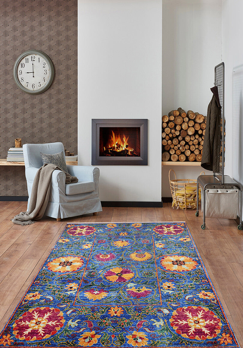 A cozy living room with the Summer Bouquet - Washable Rug - JR1358, perfect for high-traffic areas, a light blue armchair with a beige throw, wall-mounted clock, modern fireplace, and stacked firewood beside a metal bench.