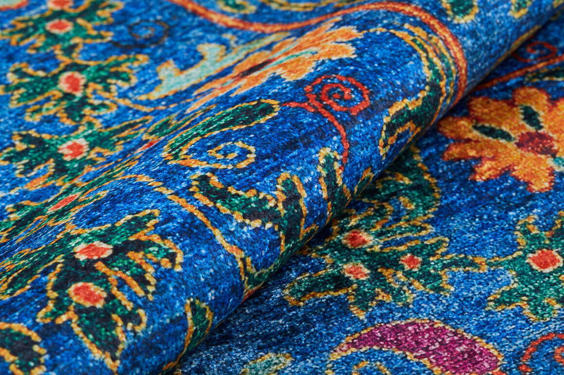 A close-up of the Summer Bouquet - Washable Rug - JR1358 shows its vibrant blue base with intricate floral and swirl designs in yellow, orange, green, and red; part of the washable rug is folded—ideal for high-traffic areas.