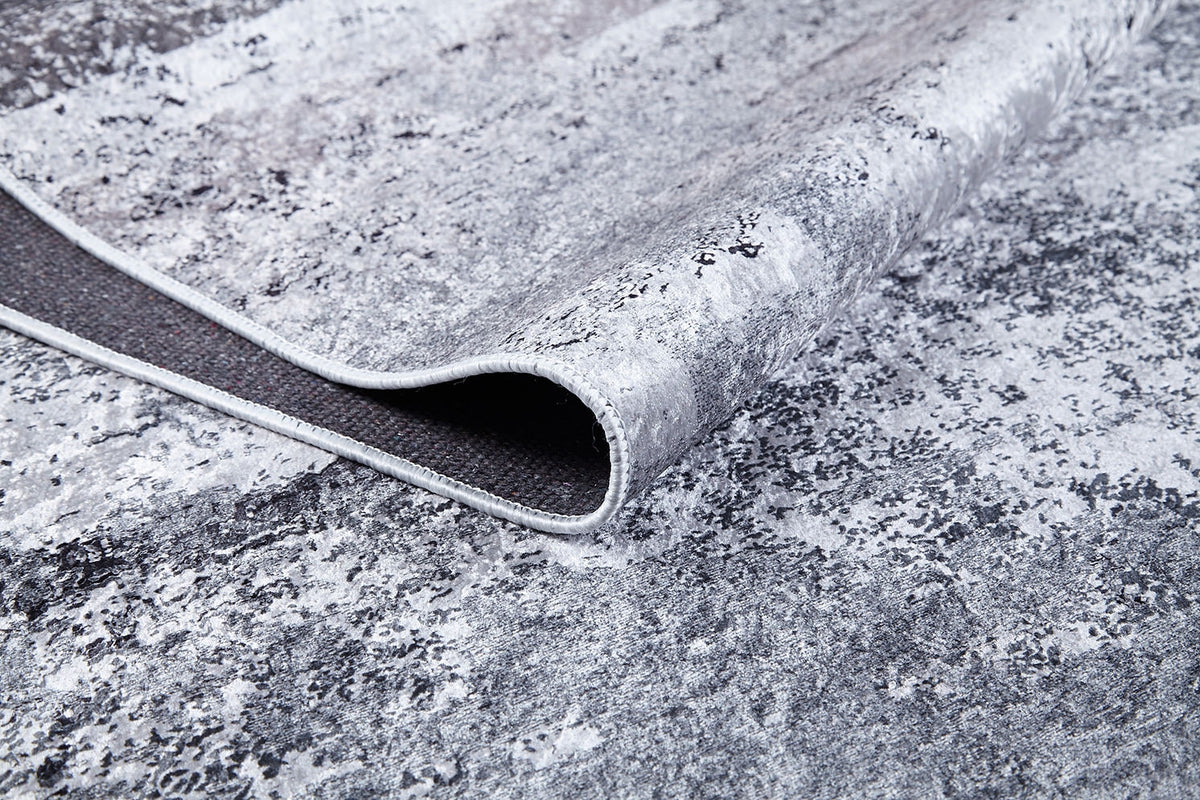 A close-up shows the corner of the Techno Flux - Washable Rug - JR1814 in gray-anthracite, slightly lifted to reveal its dark backing and edge stitching. The marbled, abstract pattern makes it ideal for high-traffic areas.