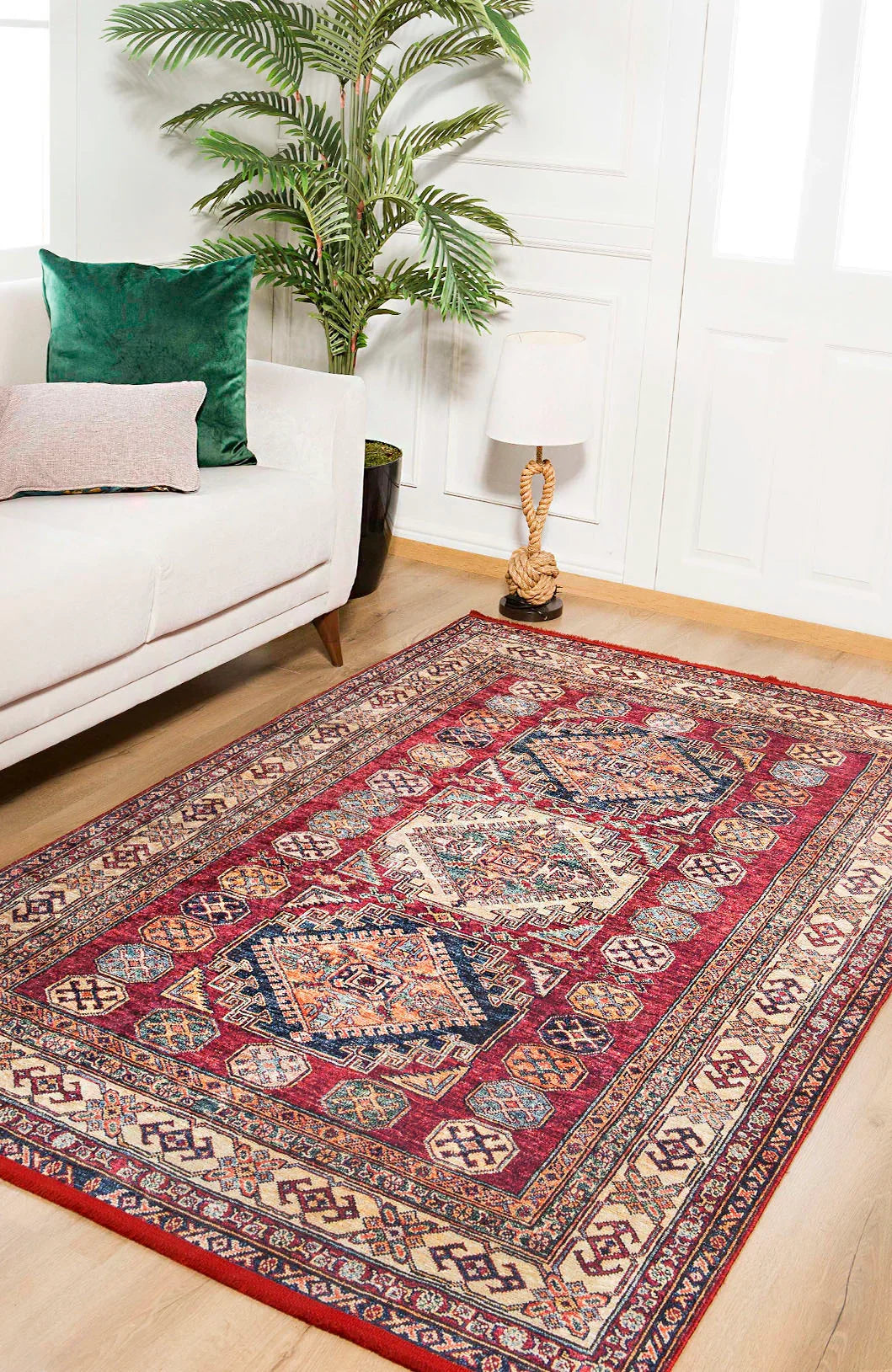 A living room with a white sofa, green and pink cushions, a potted plant, table lamp, and the Time-Honored Textures Washable Rug - JR40 in red on light wooden floors. White paneled walls and windows are in the background.