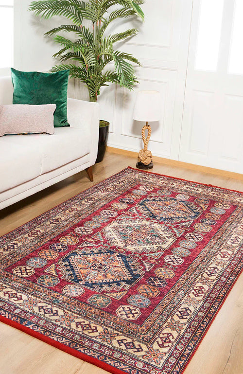 Load image 1 in gallery view. A living room with a white sofa, green and pink cushions, a potted plant, table lamp, and the Time-Honored Textures Washable Rug - JR40 in red on light wooden floors. White paneled walls and windows are in the background.