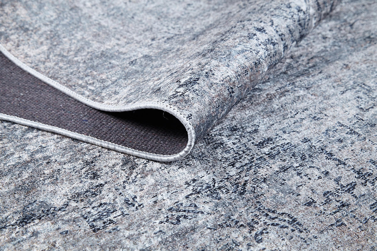 A close-up of the Timeless Scrollwork - Washable Rug - JR1774 showcases its modern gray-anthracite abstract pattern. A corner is slightly flipped, revealing the durable underside and stitched edge—ideal for high-traffic spaces.