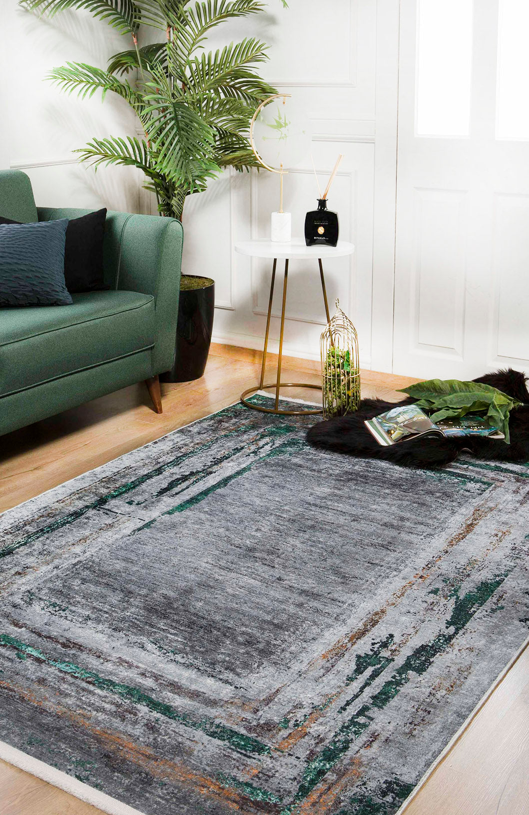 A modern living room with a green sofa, the Traditional and Classic - Washable Rug - JR1543 in gray and green, a round white side table with décor items, a tall potted plant, wooden floors, white walls, and a magazine on the floor.