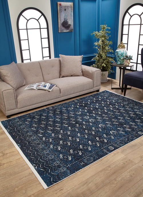 Load image 1 in gallery view. A modern living room features a beige sofa, open magazine, houseplant, small side table and chair, plus the Tribal Trail Washable Rug - JR1939 in blue—perfect for high-traffic areas. Two arched windows are set on a blue accent wall.