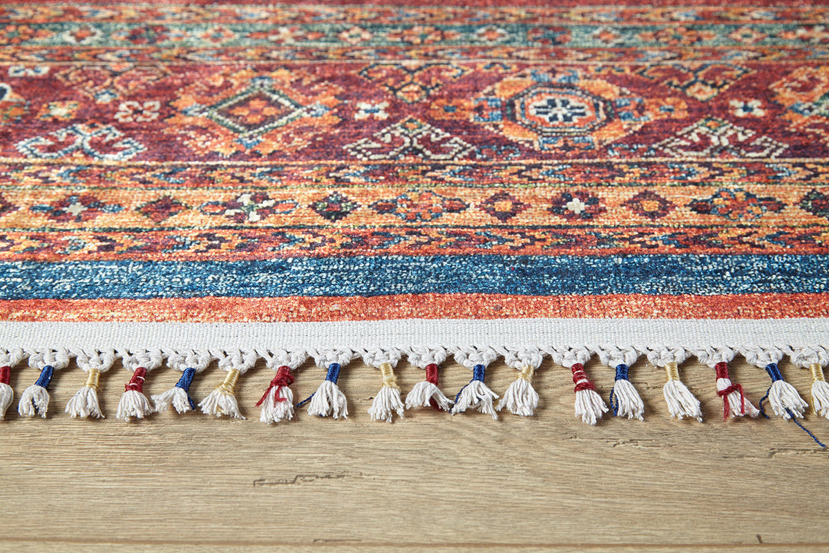 A close-up of the Tropic Thunder - Washable Rug - JR5077 shows its multicolor geometric design in red, blue, and yellow. White, red, and blue tassels line the edge. Perfect for high-traffic areas, it's shown on a light wooden floor.