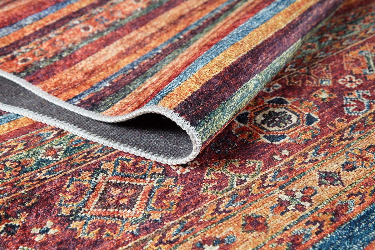 A close-up of the Tropic Thunder - Washable Rug (JR5077) shows its vibrant geometric and floral patterns in red, orange, yellow, green, and blue. A folded corner reveals the stitched edge—durable for high-traffic spaces and easily washable.