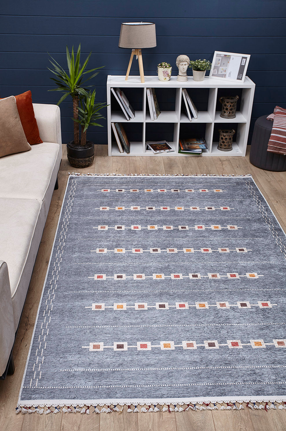 A modern living room features the Tuscan Villa - Washable Rug - JR5058 in gray-anthracite, a white cube bookshelf, books, décor, a potted plant, a beige sofa with cushions, and a table lamp against a dark blue wall—ideal for high-traffic areas.