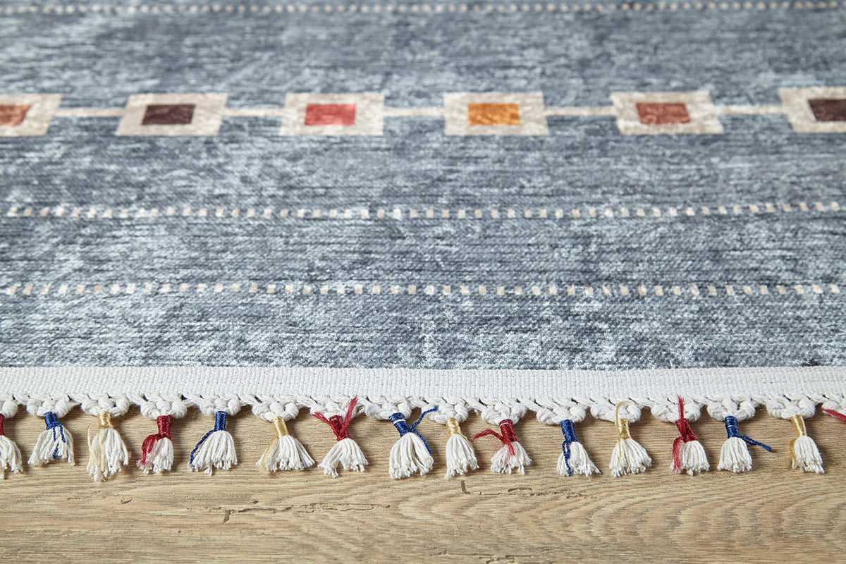 A close-up of the Tuscan Villa - Washable Rug - JR5058 shows its blue textured pattern, multicolored edge squares, and white fringe with red, blue, and yellow threads—ideal for high-traffic areas on a light wooden floor.