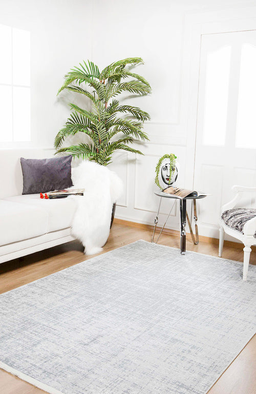 Load image 1 in gallery view. A bright, modern living room featuring a white sofa, gray pillow, faux fur throw, large potted plant, glass side table with books and a face sculpture, white armchair, and the Twilight Sky - Washable Rug - JR856 in gray-anthracite for high-traffic areas.