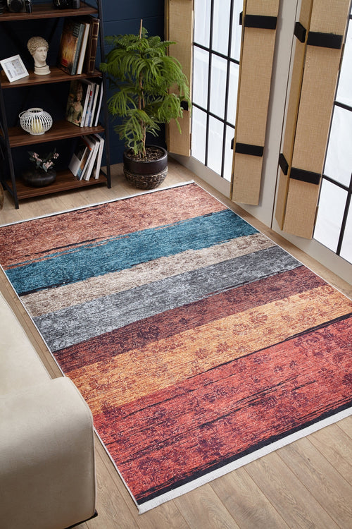 Load image 1 in gallery view. The Urban Edge - Washable Rug - JR2005, featuring multicolor horizontal stripes in blue, gray, orange, and red, adds a cozy touch to a living room with wooden floors, large bamboo-shaded windows, shelves, and a potted plant.