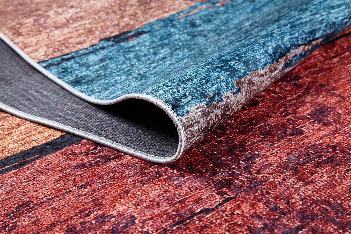A close-up of the Urban Edge - Washable Rug - JR2005 shows its textured, abstract pattern in blue, red, and brown tones with part of the flat weave folded to display the underside.
