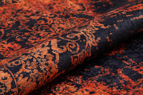 Load image 1 in gallery view. A close-up of the Vintage Glory - Washable Rug - JR1490 shows its ornate black and orange swirling and floral patterns on a textured surface. The partially rolled rug highlights its vibrant colors—ideal for high-traffic areas.