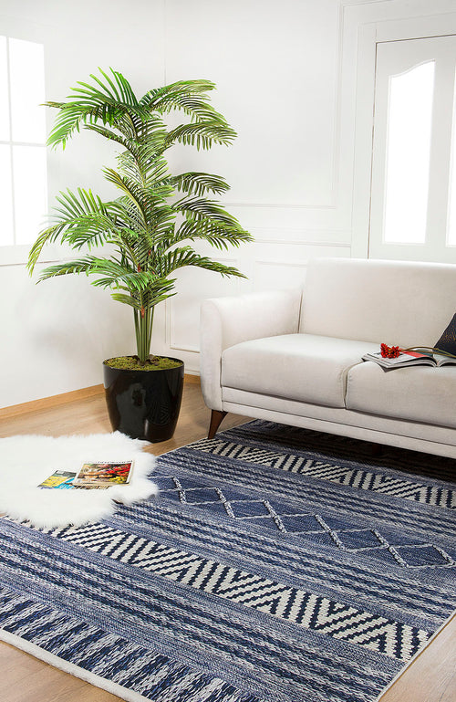 Load image 1 in gallery view. A bright living room features a white sofa, a large plant, and the Wavy Wonders - Washable Rug - JR941 in blue—perfect for high-traffic areas—with a fluffy white rug and magazines on light wooden floors by large windows.