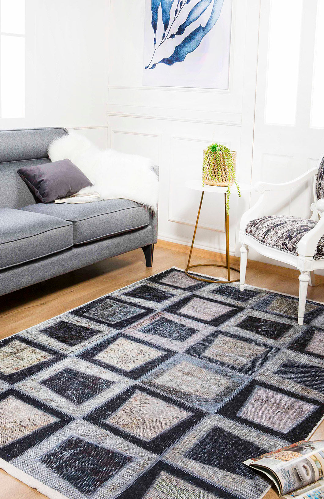 A modern living room with a gray sofa, white armchair, small round side table with a plant, abstract artwork, and the Windowpane Wonder - Washable Rug - JR184 in a geometric pattern on the wooden floor.