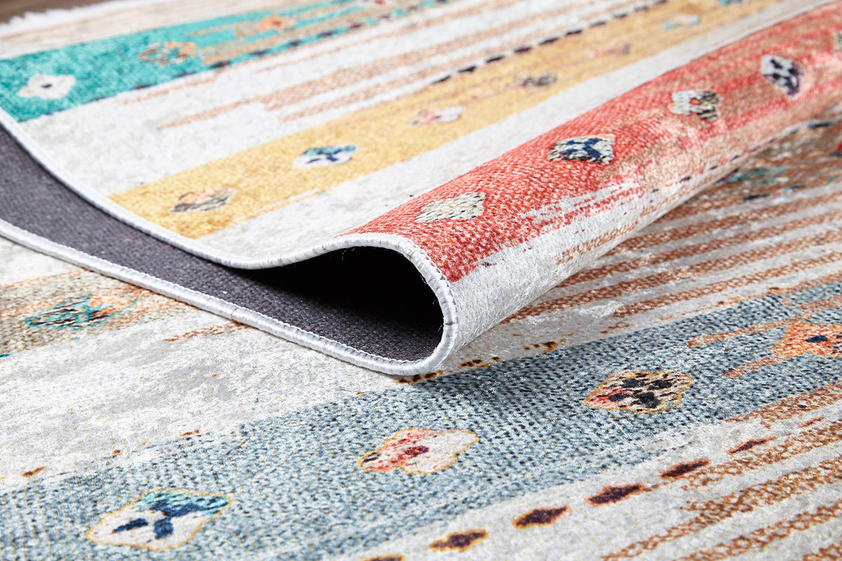 A close-up of the Nomad's Nostalgia - Washable Rug (JR1669) shows vibrant stripes and geometric shapes, with a folded corner revealing its dark underside—ideal for brightening high-traffic spaces.