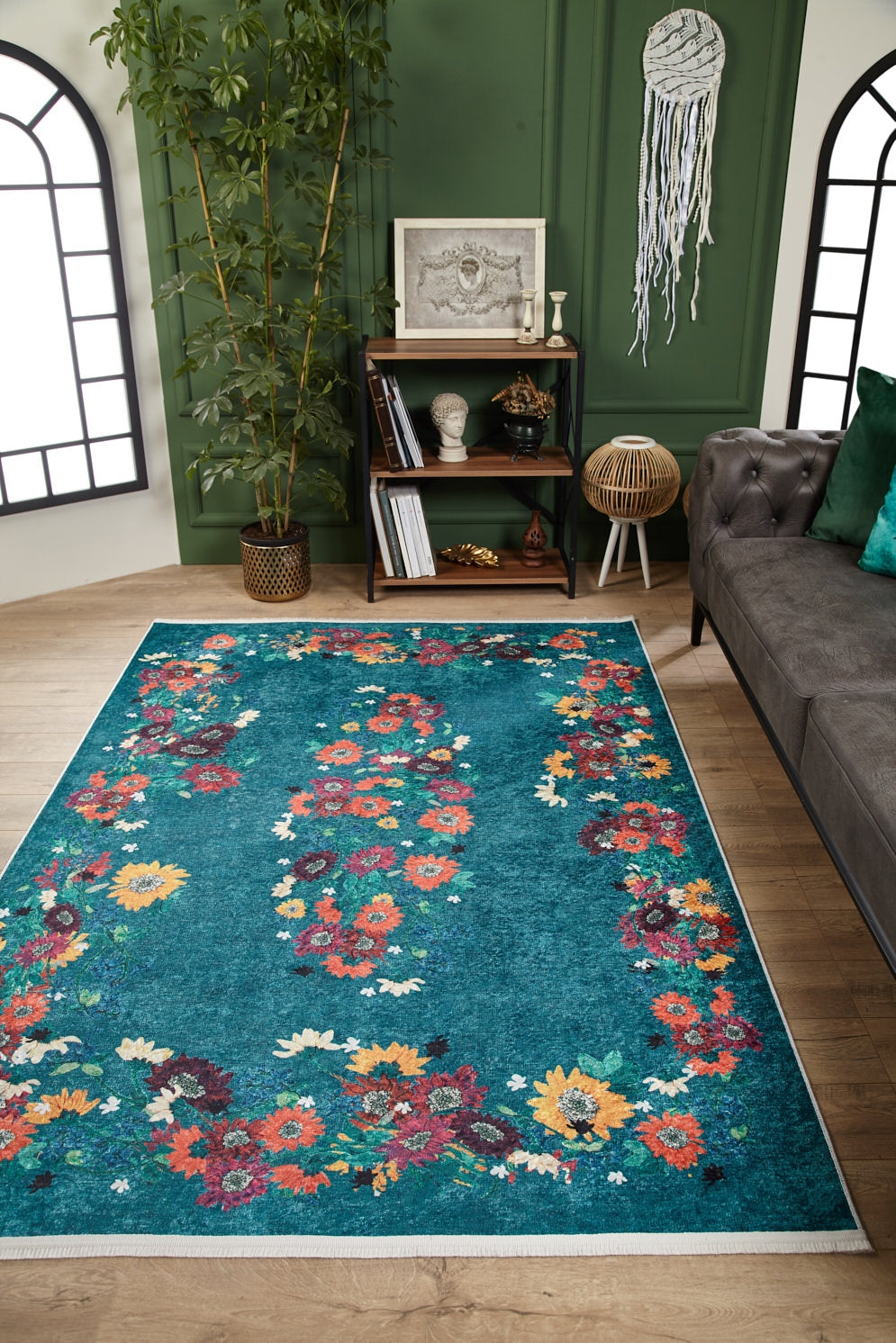 A living room features the Nature's Tapestry - Washable Rug - JR1674 in teal floral, a gray sofa, bookshelf, green accent wall, large potted plant, and framed artwork with a hanging macrame dreamcatcher—ideal for high-traffic spaces.