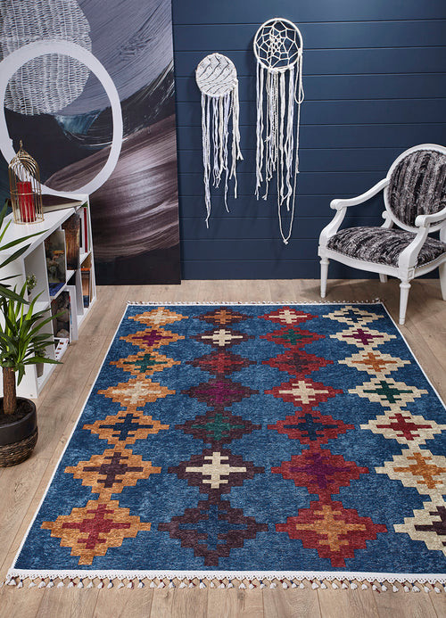 Load image 1 in gallery view. The Nomad's Mirage - Washable Rug - JR5032, featuring blue, red, yellow, and cream geometric patterns, is laid on a light wood floor near a white chair, indoor plant, bookshelf with books, and two white macramé wall hangings.