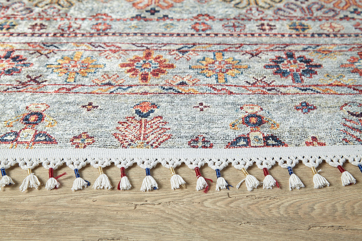 The Nomad's Nest - Washable Rug - JR5088 features floral and geometric patterns in red, blue, yellow, and orange with cream fringe tassels. Shown on a wooden floor, it's perfect for high-traffic areas and easy to clean.