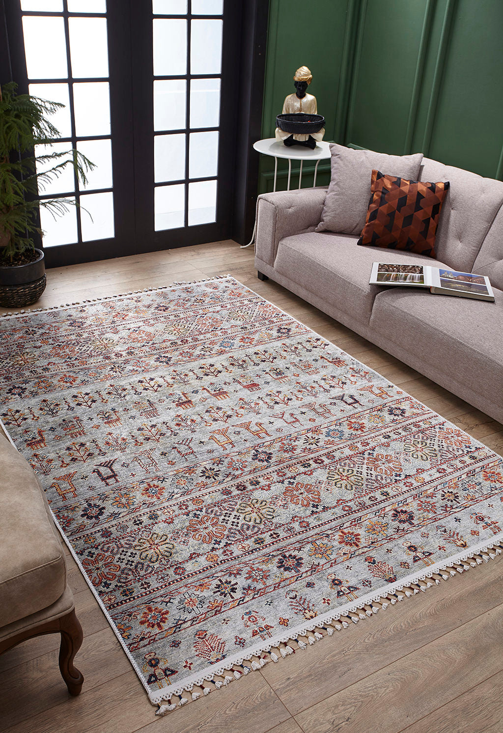 A living room features the Nomad's Nest - Washable Rug - JR5088, perfect for high-traffic areas, paired with a light gray sofa, patterned pillow, small round side table with decor, potted plant, and large black-framed windows filling the space with light.